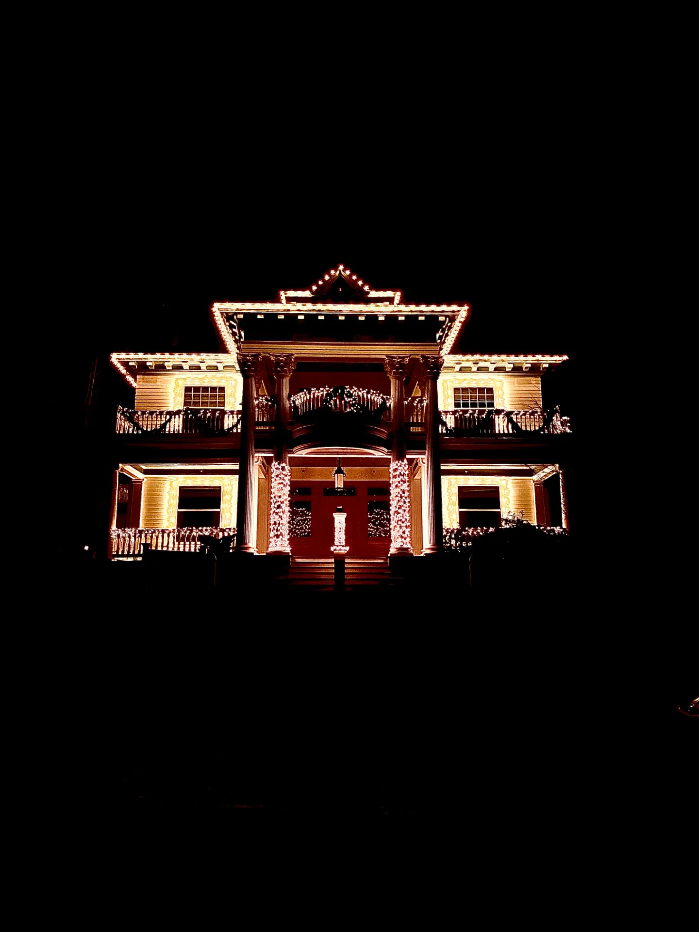 Colonial-style home glowing with warm white C9 lights along every roofline, balcony, and column against a black night sky